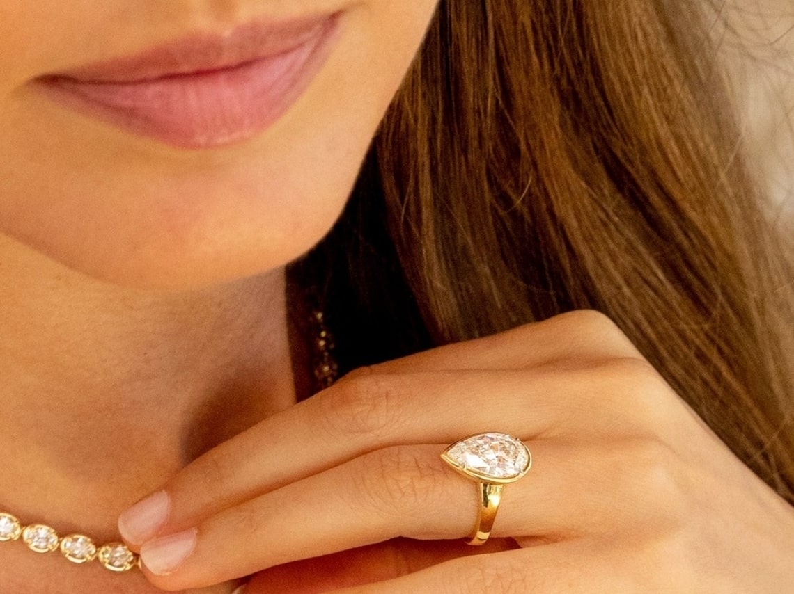 A woman smiling while wearing a yellow gold pear shaped bezel set diamond solitaire engagement ring and touching a yellow gold and diamond collar necklace.