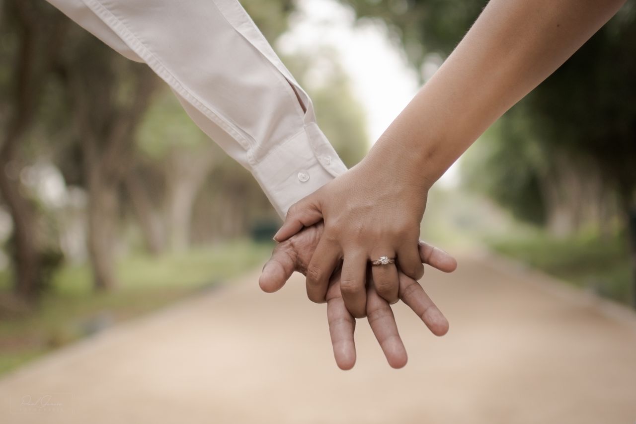 A man and woman holding hands on a path through a park with the focus on the diamond engagement ring freshly added to her hand after the proposal. 